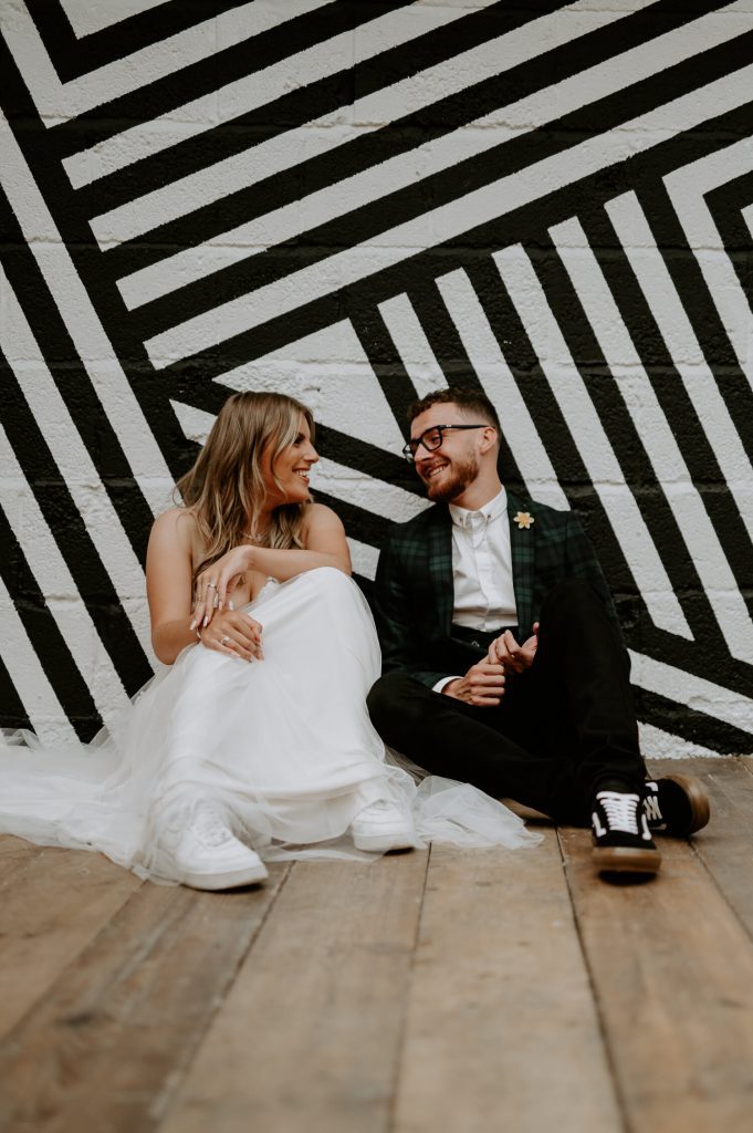 A bride and groom sit next to a black and white mural at the shack revolution. The bride is wearing a wearing dress and nike air force 1 trainers. The groom is wearing a tartan suit and back and white vans authentic trainers.
