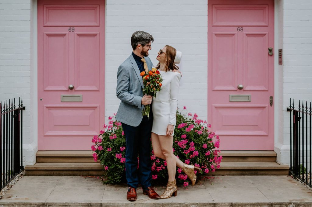 A bride wearing a short wedding dress stars into her grooms eyes whilst standing outside the pink doors of Turner Studios in Chelsea, London.