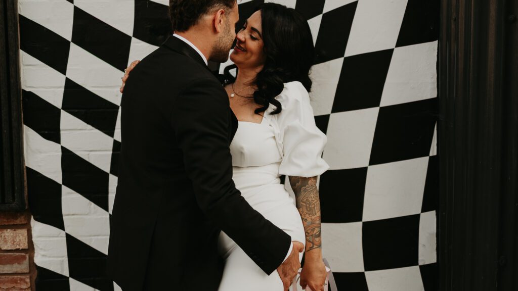 A bride and groom kiss in front of the black and white checked mural at The Shack Revolution. 