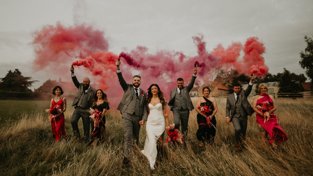 A Family Photo at a Wedding of a bride and groom with their wedding party walking through a field with pink and orange smoke grenades.