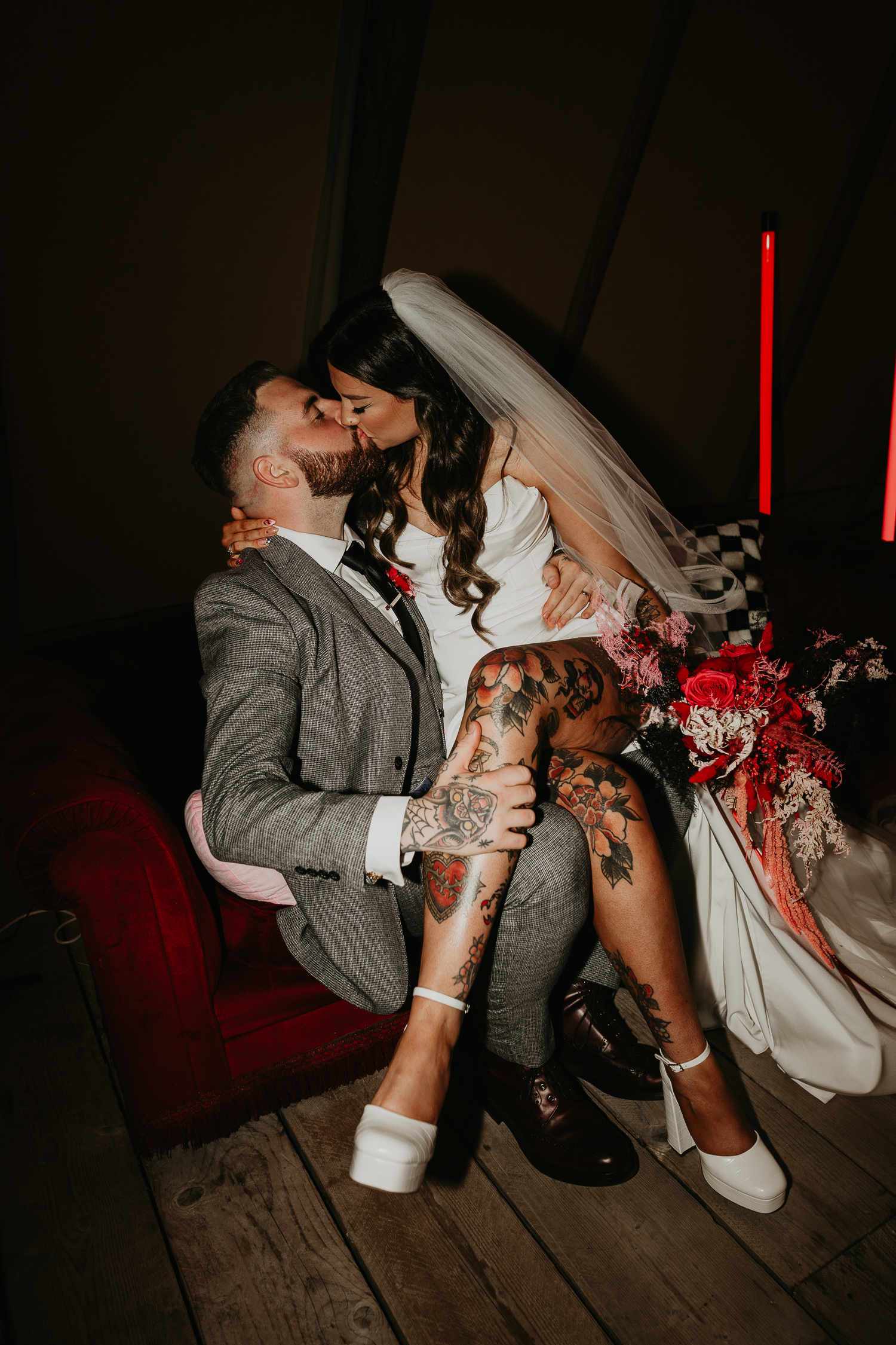A bride wearing a slip wedding dress with her tattooed leg showing is sat on her grooms lap kissing him.