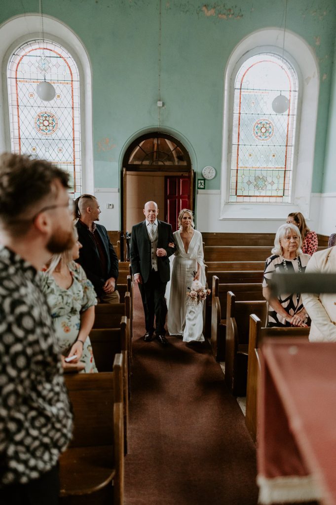 A Bride and her Father walk into a vegas style church in Bristol as the groom looks down the aisle at them.