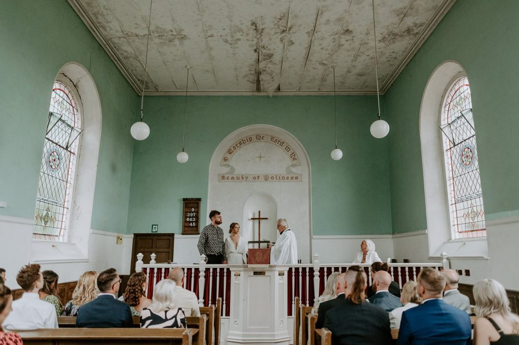 A bride and groom stand at the front of a small vegas style church in Bristol with a vicar as he marries them.