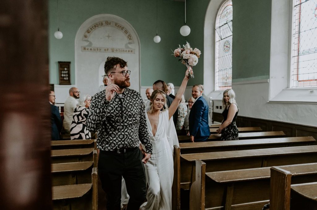 A bride raises her bouquet as her and her groom walk out of a small church in Bristol.