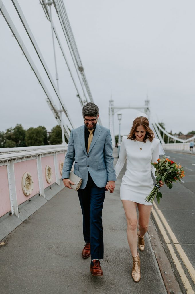 A bride and groom walk over Albert Bridge near Chelsea. The bride is wearing a short wedding dress and has a vintage veil and hat combo.