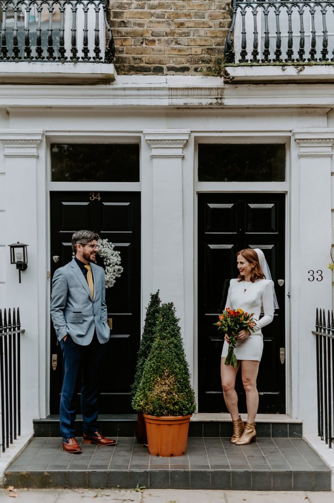 A bride wearing a short wedding dress and a groom wearing a blue jacket stand outside two black doors in Chelsea London.