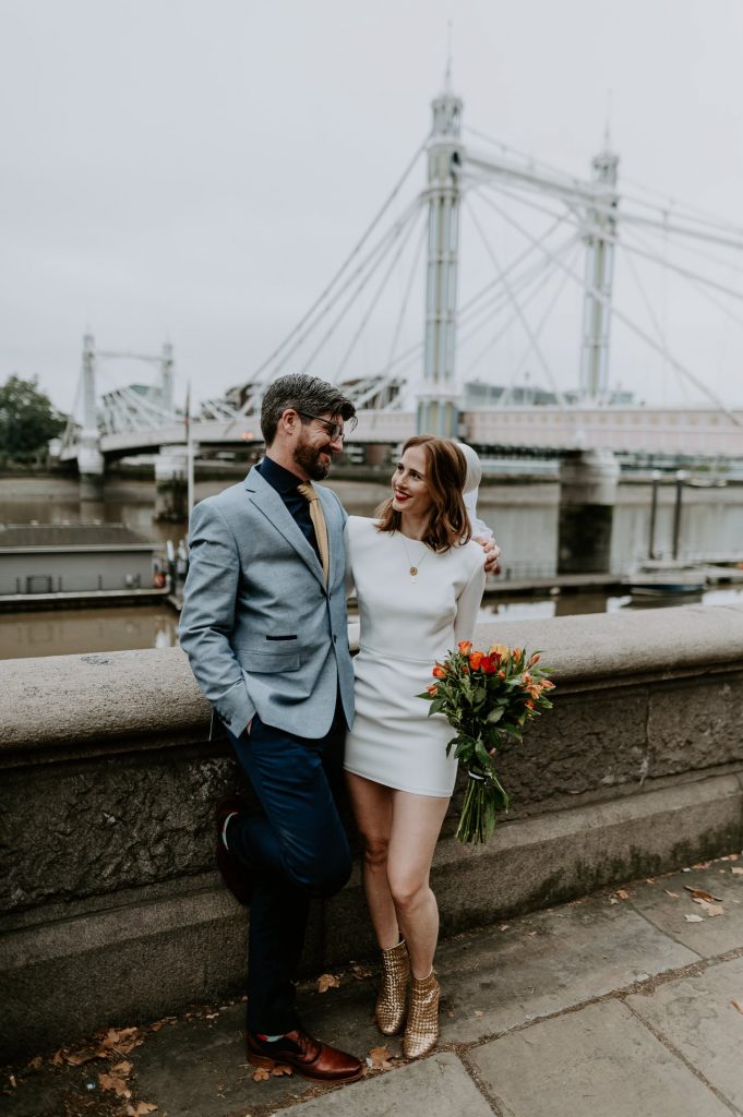 A Bride and Groom stand on the bank of the River Thames with Albert Bridge behind them.