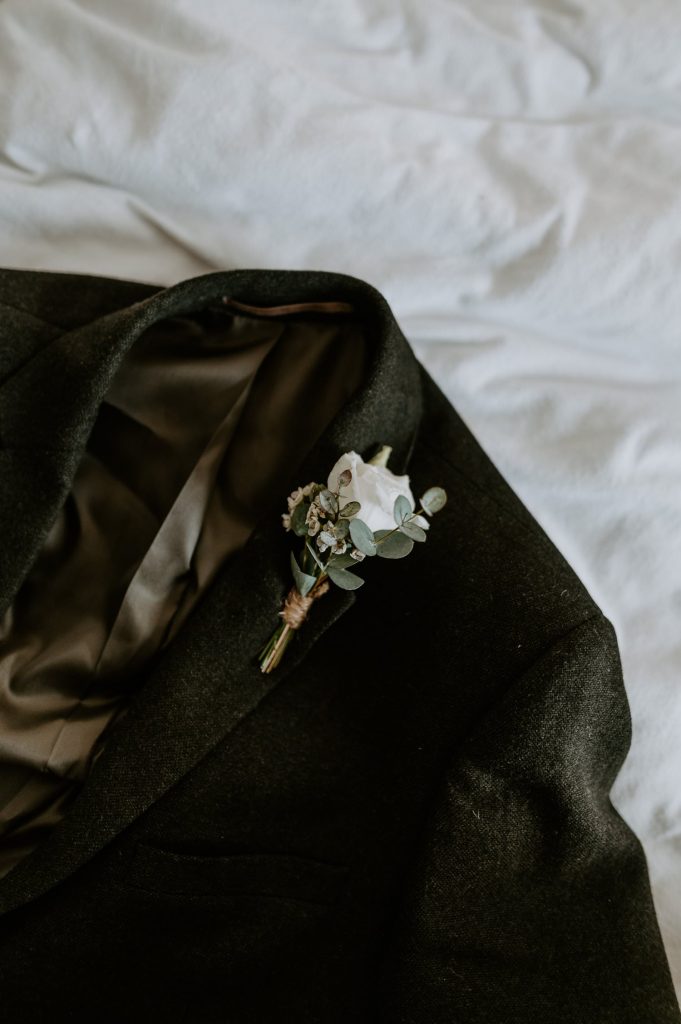 Grooms green blazer lays on bed with a white button hole pinned on.
