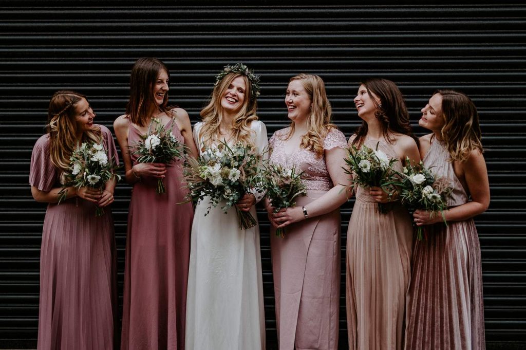 A bride and her bridesmaids laugh in front of a black shutter at Clapton Country Club.