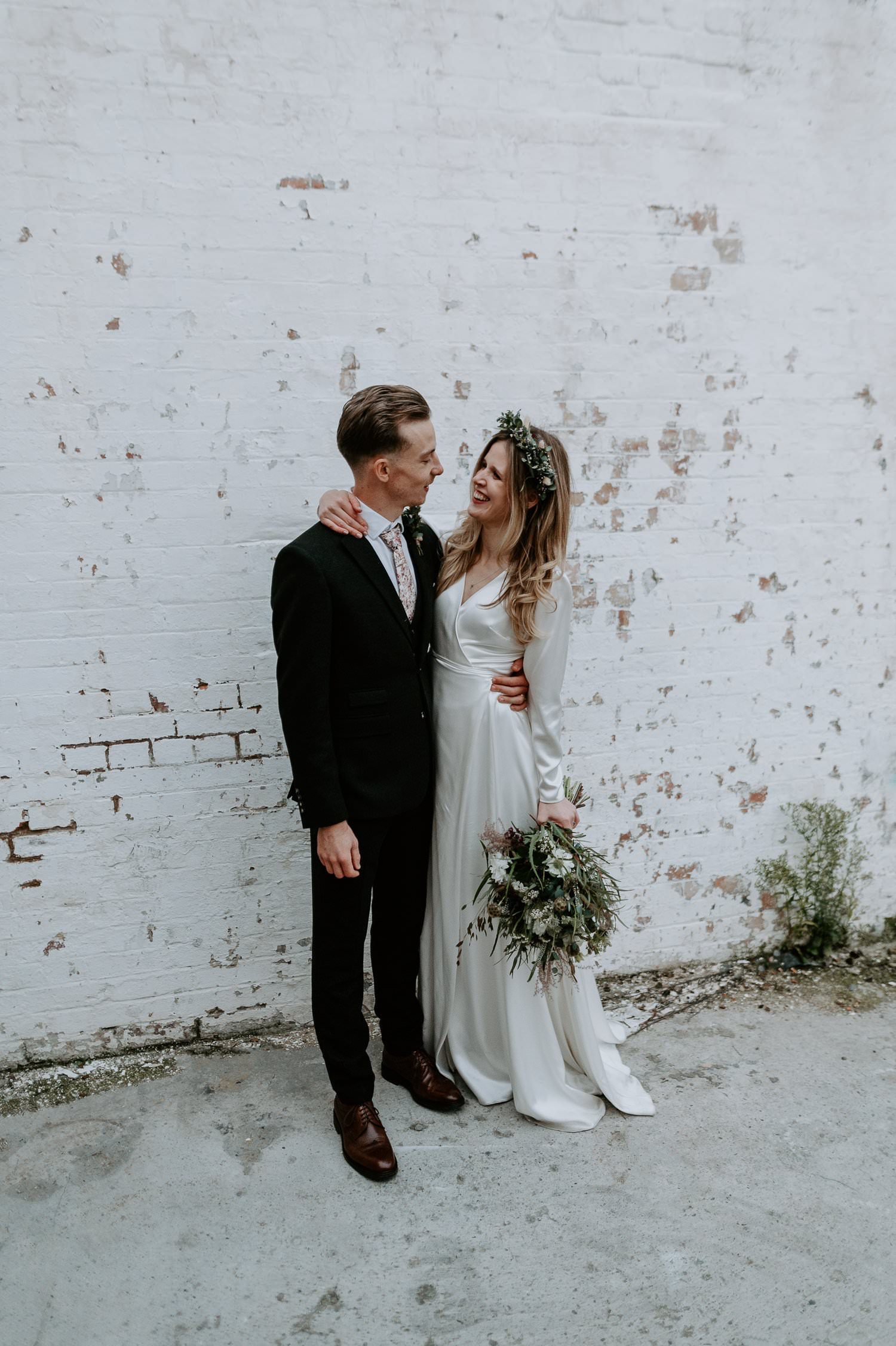 A couple on their wedding day stand by a white brick wall at Clapton Country Club. The bride is dressed has a boho dress and boho wedding flowers.
