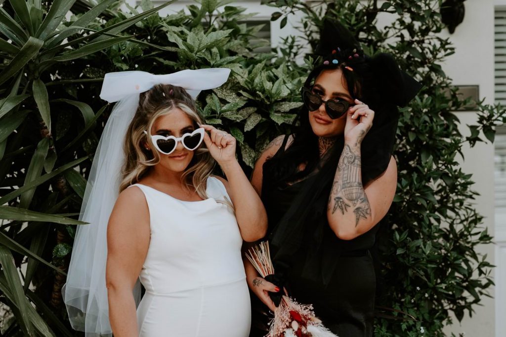 Two brides tip their heart shaped glasses towards the camera.