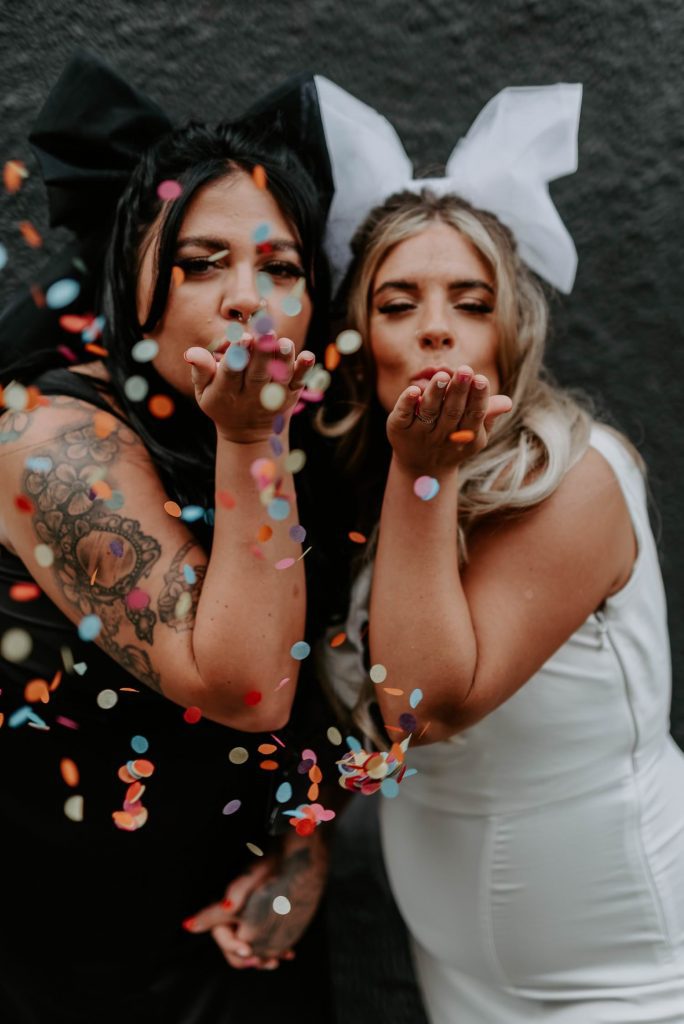 Two same sex brides blow confetti at the camera lens.