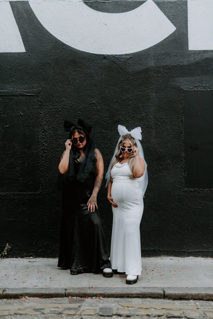 Two same sex brides stand in front of a black wall in Shoreditch and tip their heart shaped glasses.