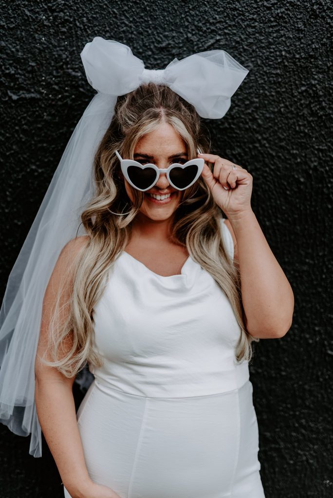 A bride stands in front of a black wall in Shoreditch and tips her heart shaped glasses.