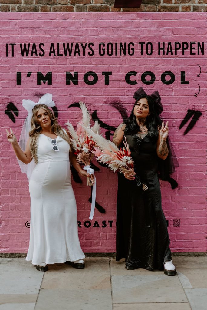 Two same sex brides stand in front of a pink wall in Shoreditch.