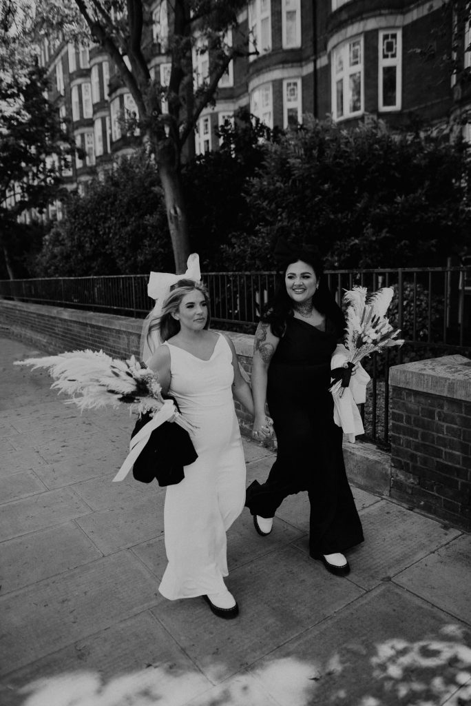 A black and white photo of two brides walking to their ceremony at Old Marylebone Town Hall in London.