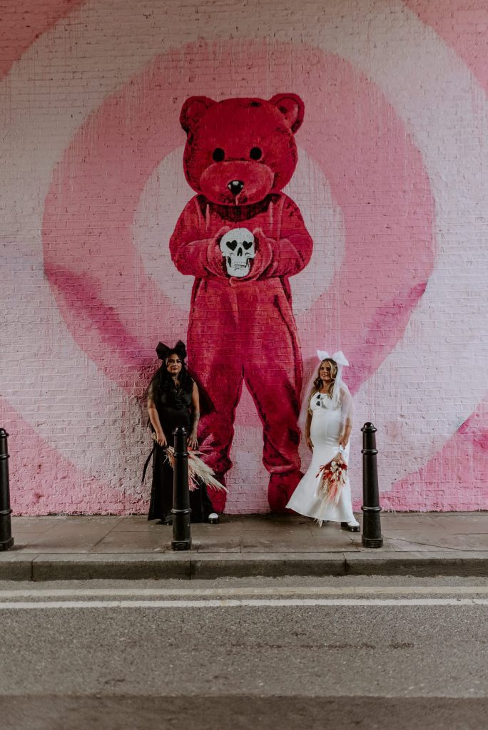 A same sex couple stand in front of a pink beard mural outside Shoreditch Studios in London on their wedding day.