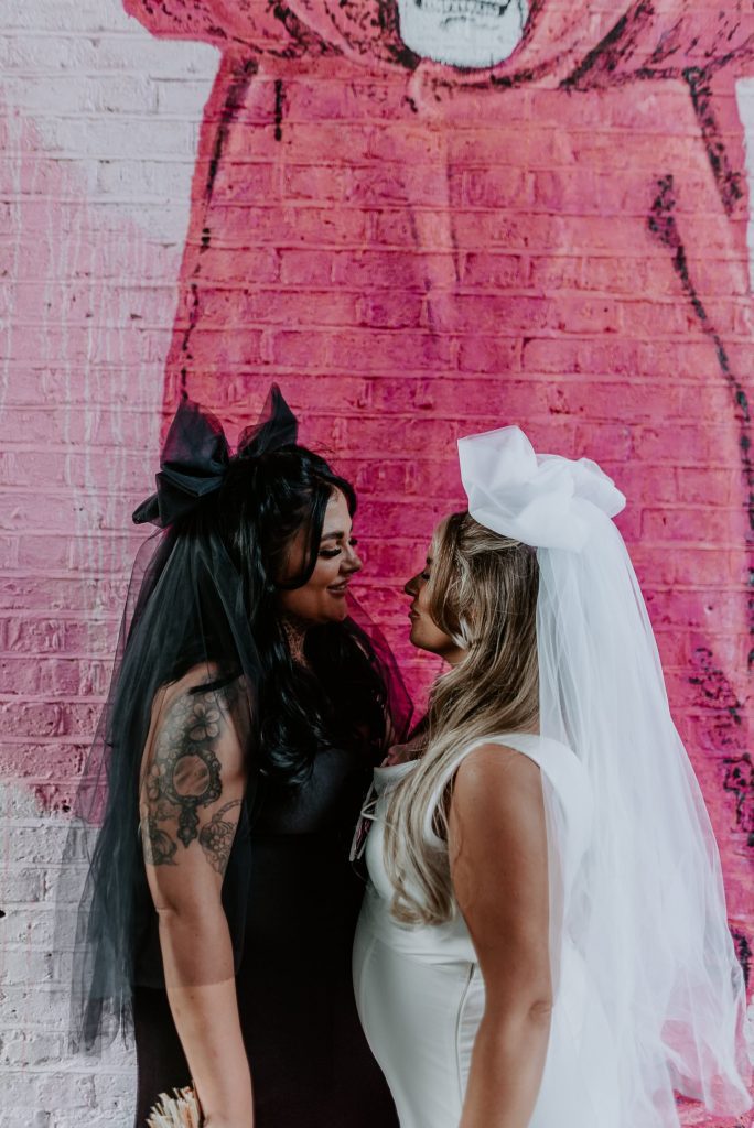 A same sex couple stand in front of a pink beard mural outside Shoreditch Studios in London on their wedding day.
