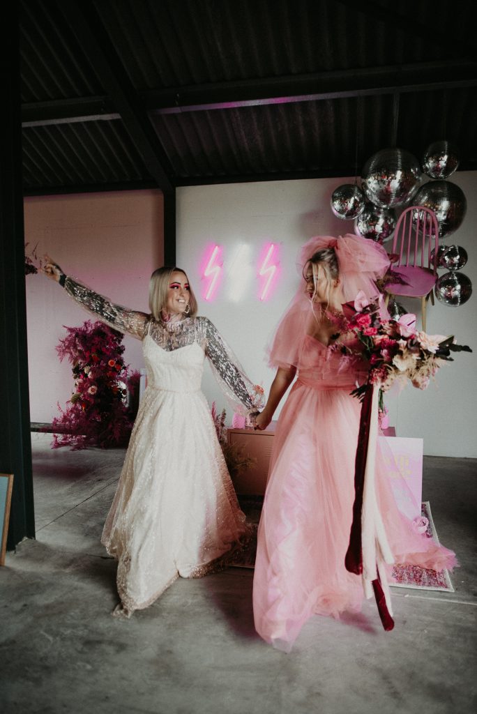 Two Brides walk out of the giraffe shed with their arms up, one is wearing a disco ball dress and the other is wearing a pink dress and holding a giant pink bouquet. You can see the pink and white thunderbolt neons of the giraffe shed in the background.