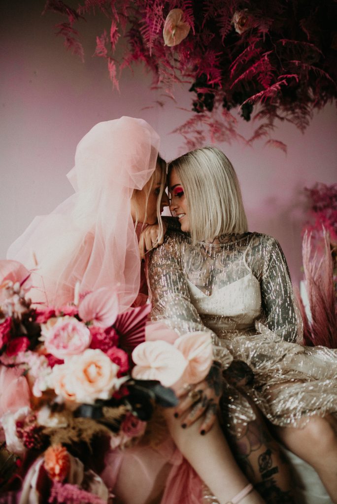 Two brides sit on a sofa at the giraffe shed, you can see they leg tattoos and shoes. One is wearing a pink wedding dress and the other is wearing a disco ball style dress.