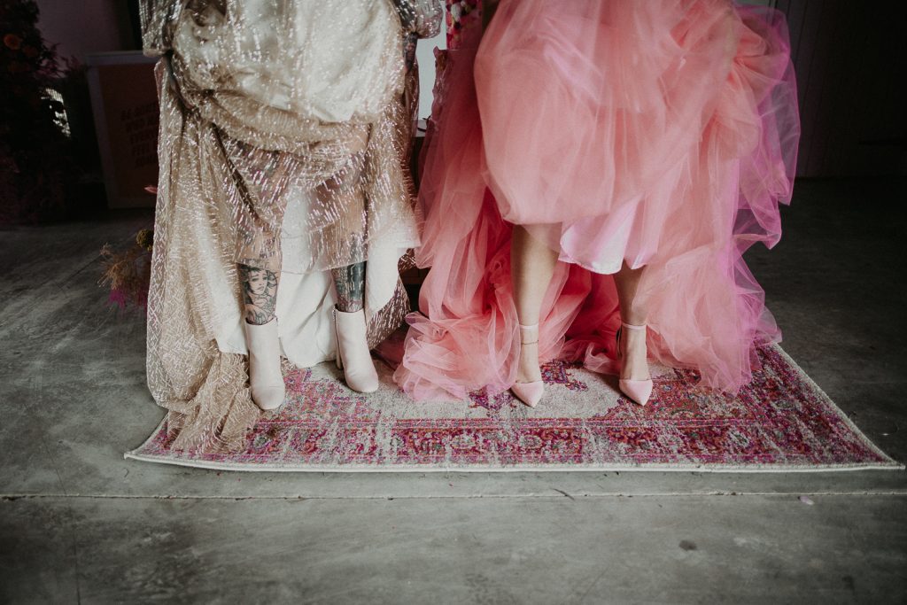 Two brides show their wedding footwear, one is wearing white boots and the other is wearing pink heels. The concrete floor of the giraffe shed contrasts with their colours.