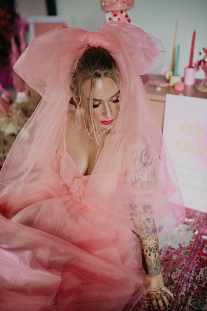A bride wearing a pink dress and veil sits on the floor at the giraffe shed. She is surrounded by pink styling as the concrete floor os the industrial wedding venue contrasts with her pink dress.