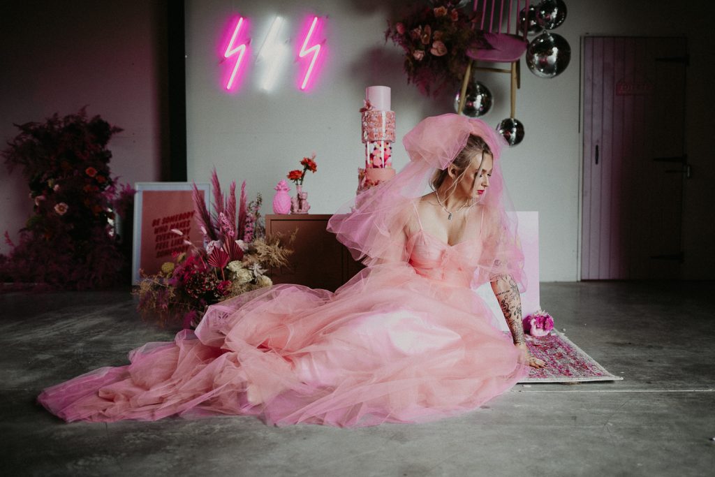 A colourful wedding shoot, a bride wearing a pink wedding dress sits on the floor. The is a pi pink thunderbolt neon in the background with a pink wedding cake.