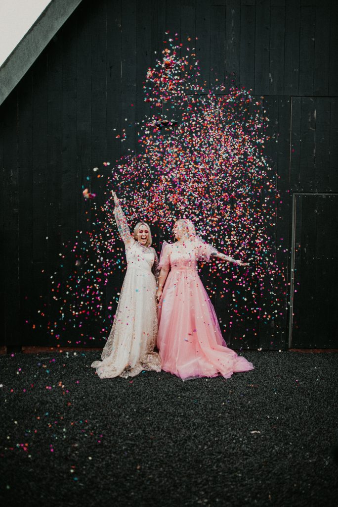 Our same sex couple throw colourful confetti outside the giraffe shed. the confetti and the girls pinks and white dresses contrast against the industrial black barn of the wedding venue.