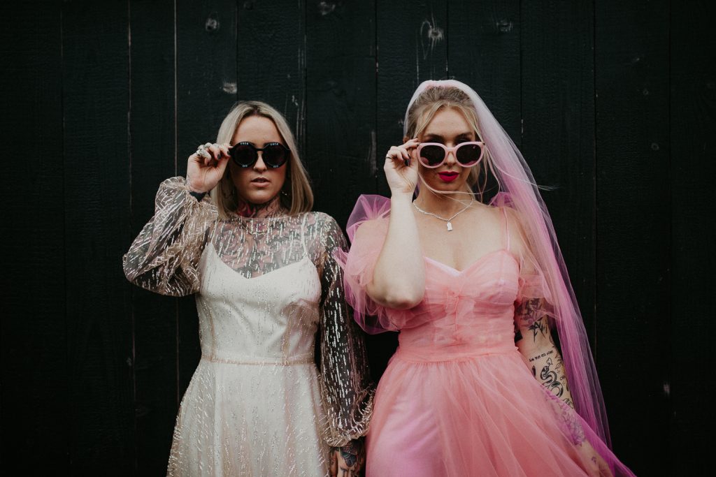 Two brides getting married at the giraffe shed are in sun glasses. One bride is wearing a pink wedding dress and the other is wearing an E&W Couture dress that looks like a disco ball.