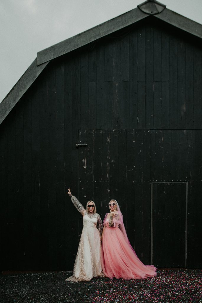 Two brides stand out the front of the giraffe shed, one bride is wearing a hot pink wedding dress and the other is wearing a white disco ball dress. They both contrast with the industrial wedding venues black barn door.