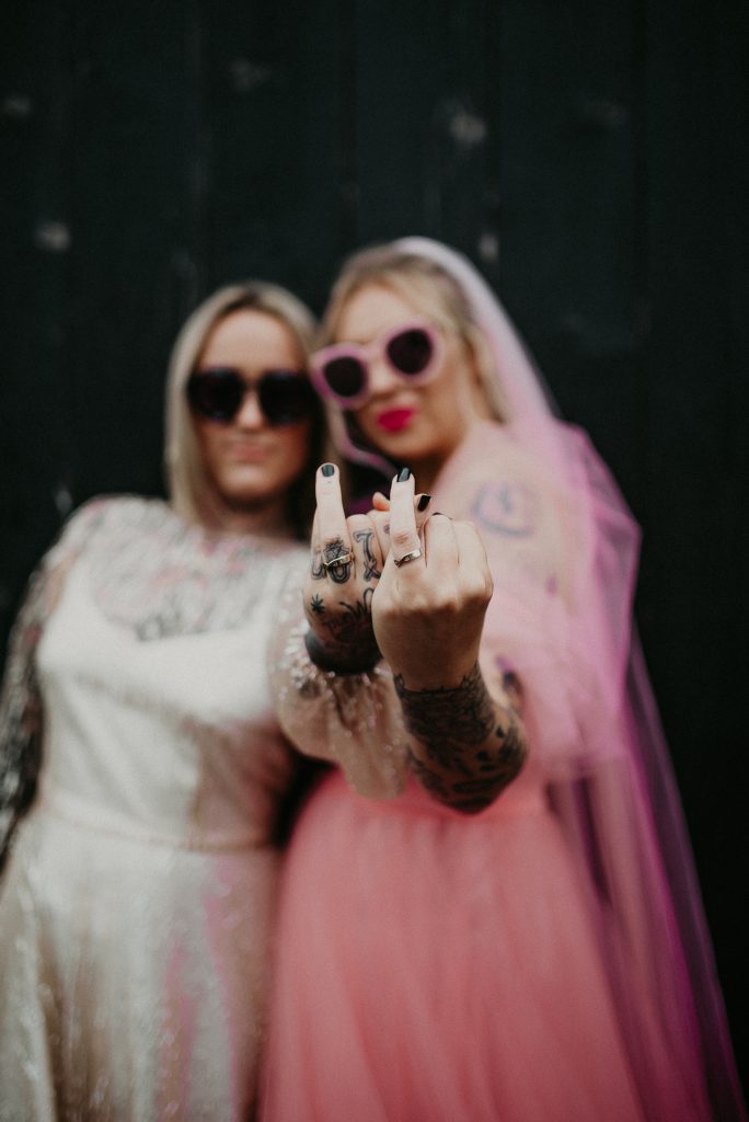 Two brides stand out the front of the giraffe shed showing their wedding rings, one bride is wearing a hot pink wedding dress and the other is wearing a white disco ball dress. They both contrast with the industrial wedding venues black barn door.