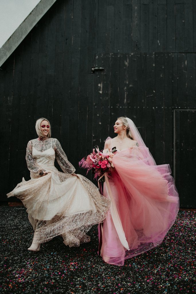 Two brides throw their dresses around outside the giraffe shed. Their dresses both contrast with the industrial wedding venues black barn door.