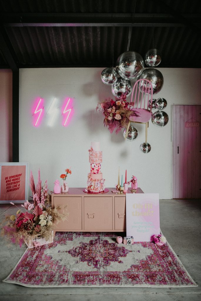 A pink wedding cake sat on a peach locker s surrounded by pink flowers and disco balls. Luna and the Lane's styling at The Giraffe Shed.