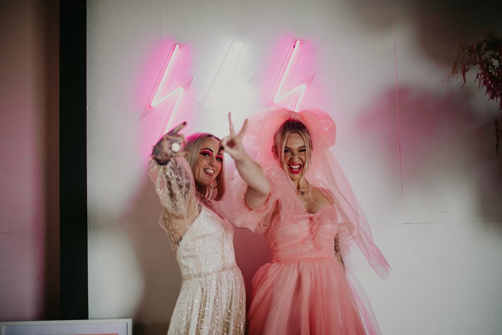 Two women at the giraffe shed, one wearing a pink wedding dress and the other wearing a disco ball wedding dress throw a peace sign.