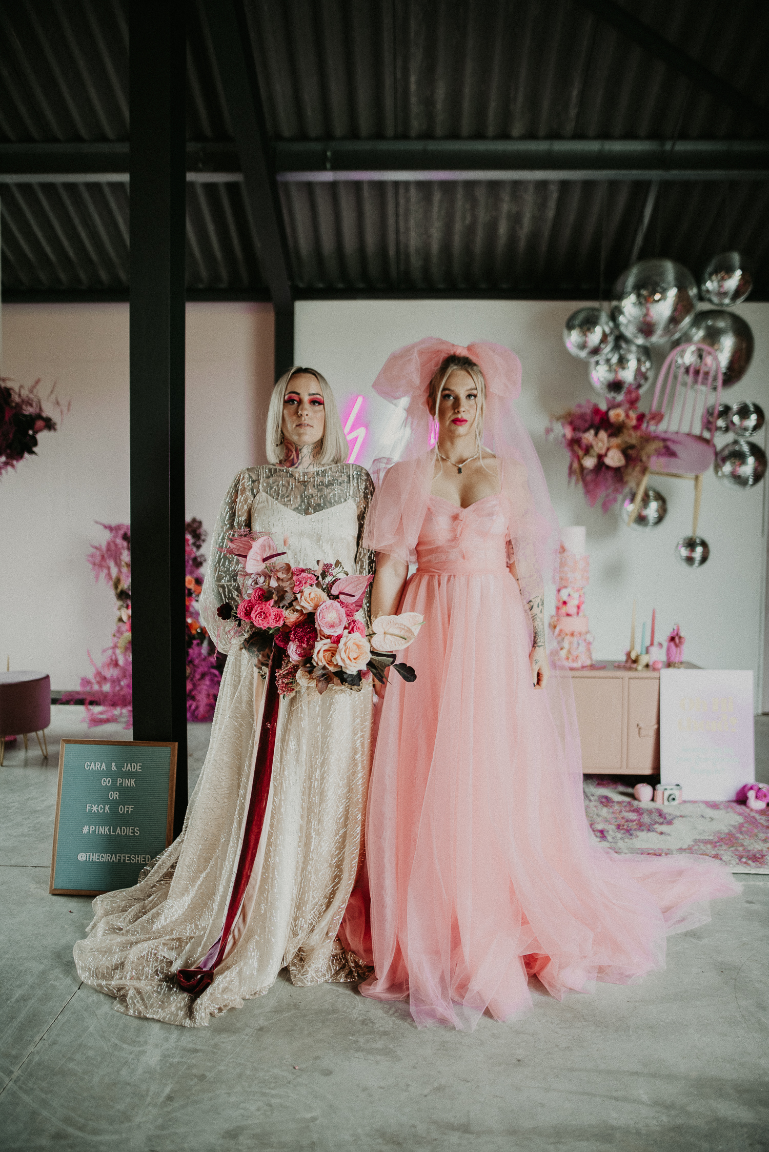 Same Sex wedding at the giraffe shed. Two brides in dresses, one pink and one white pose for a photo in front of a pink neon at the giraffe shed.