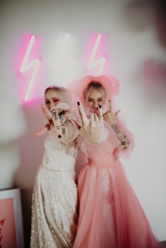 Two women at the giraffe shed, one wearing a pink wedding dress and the other wearing a disco ball wedding dress throw the rock hands.
