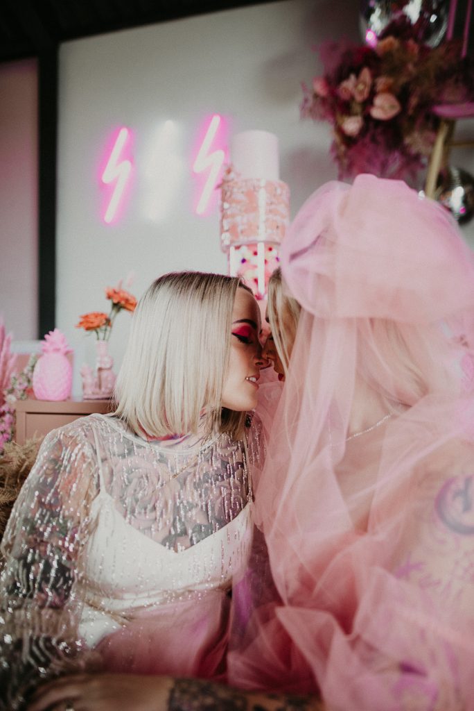 Two brides sat on the floor of the giraffe shed, in the background you can see a pink cake and pink and white neons.