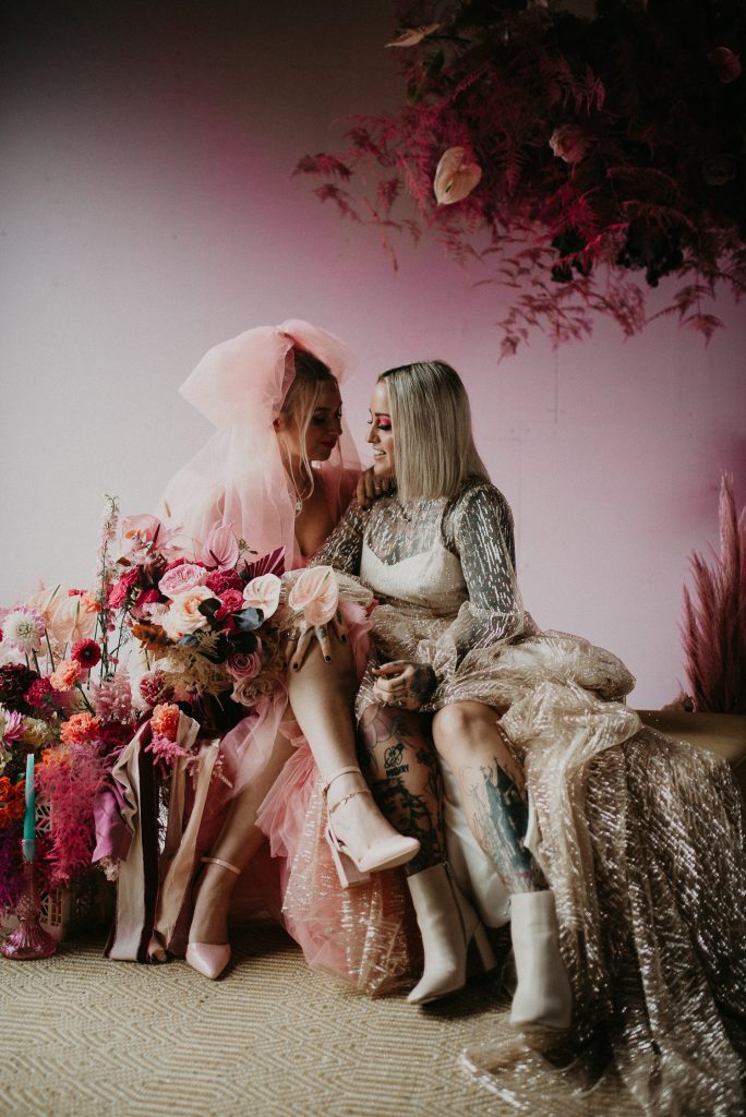 Two brides are sitting on a sofa at The Giraffe Shed. Surrounding them are huge pink flowers. One bride is wearing a pink wedding dress and the other is wearing a wedding dress that looks like a disco ball.