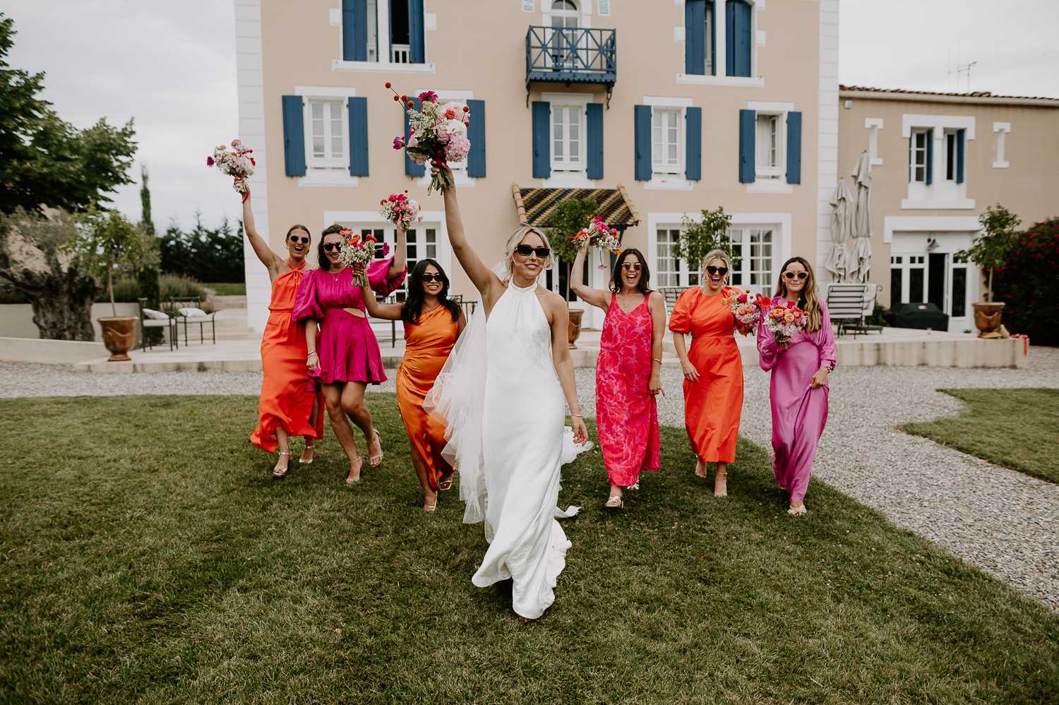 A bride and her bridesmaids throw the bouquets in the air as they walk in a line. The bouquets are all different shades of pinks and oranges and her bridesmaids wore pink and orange dresses that were all different styles and had their own pink bouquets.