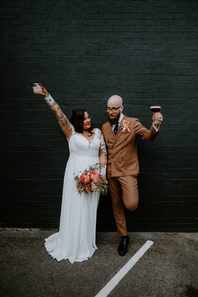 A bride and groom stand against a black wall at Five Four Studios with their glasses in the air. The groom is wearing a tobacco brown suit and you can see the brides tattoos through her dress.