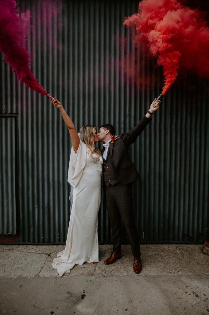 A bride and groom kiss in front of a black shutter door whilst holding pink and red smoke grenades.