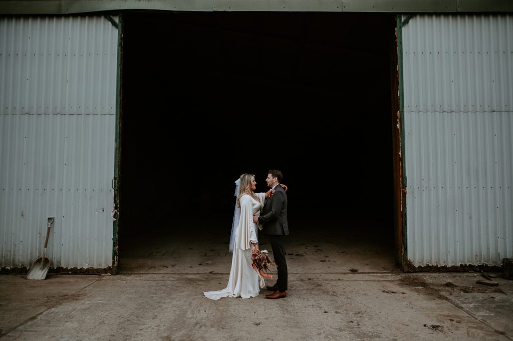 A bride stands with her hand on her grooms shoulder outside a giant barn door.