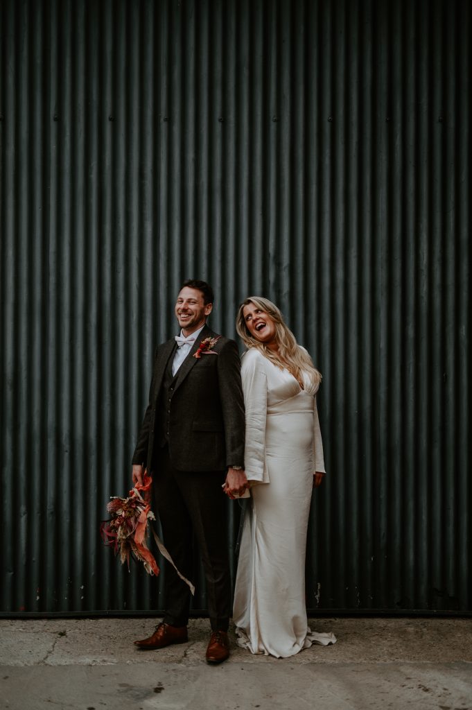 A bride and groom stand back to back and laugh in front of black shutter door.