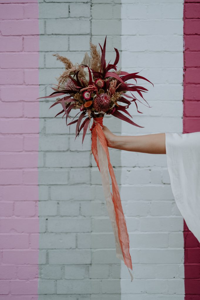 A dried bouquet of pink and purple flowers being held up in front of a pink wall.