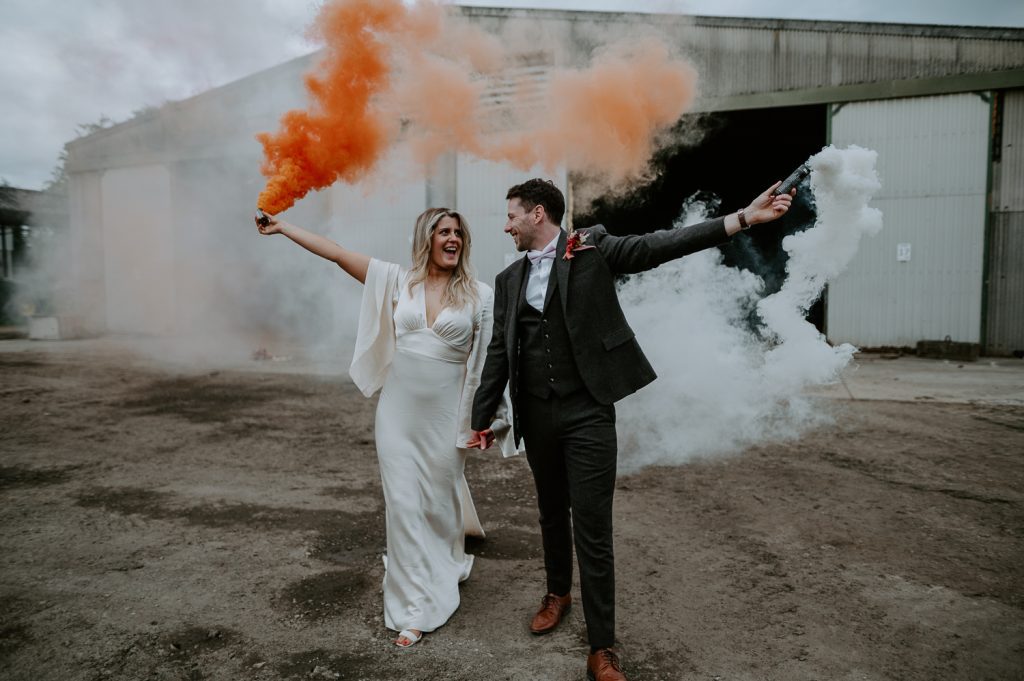 A bride and groom walk away from a barn whilst laughing at each other whilst holding white and orange smoke grenades.