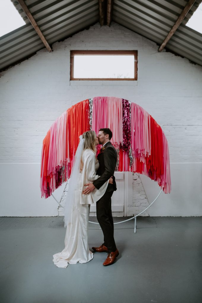 A bride and groom kiss in front of their pink streamer back drop dusting their wedding ceremony at White Syke Fields in York.