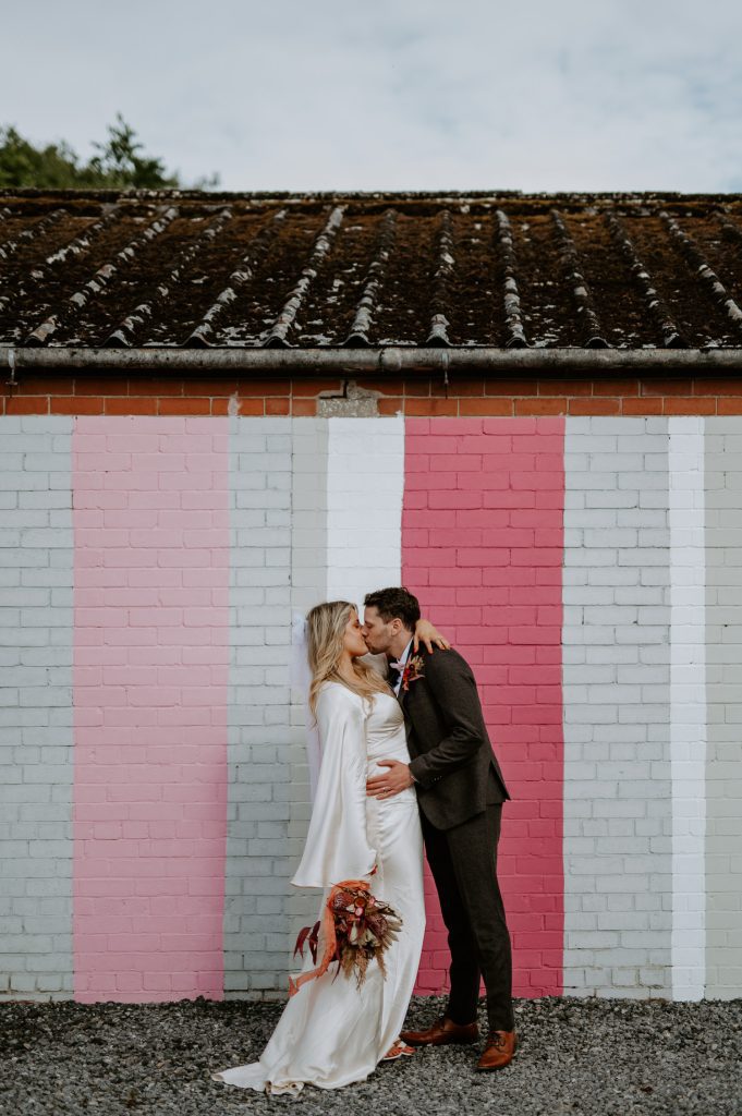 A bride and groom kiss in front of the pink wall at White Syke Fields wedding venue in York.