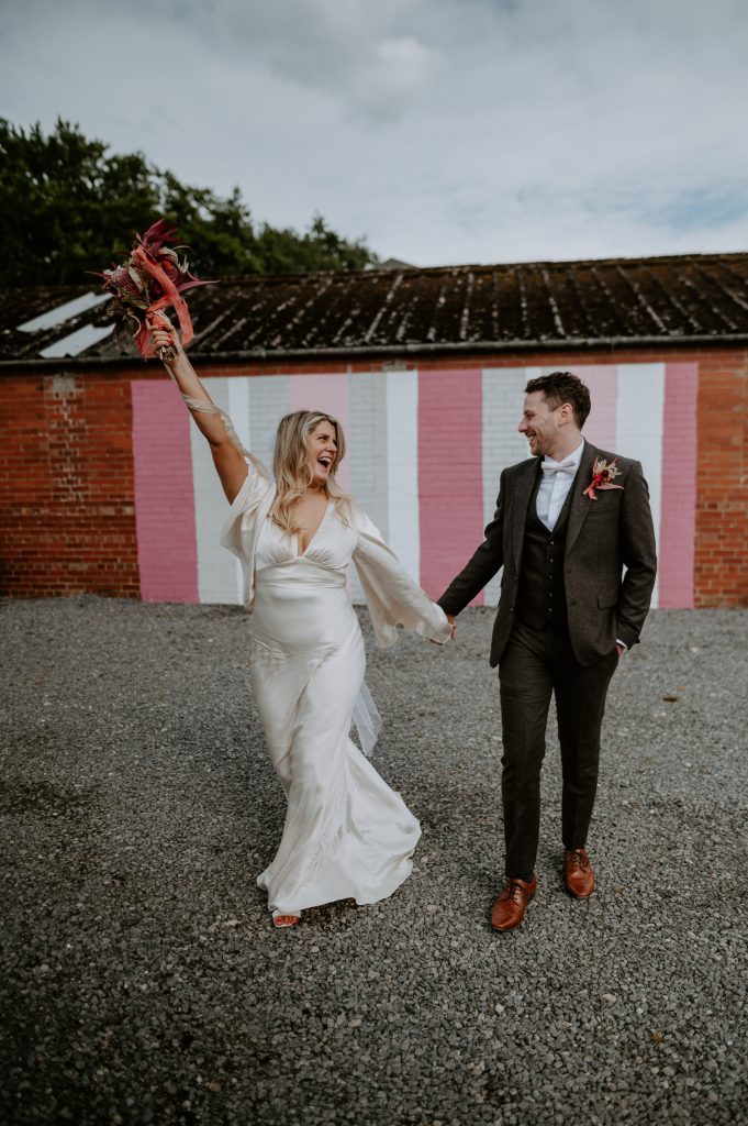 A bride throws her bouquet in the air and smiles at a groom whilst walking.