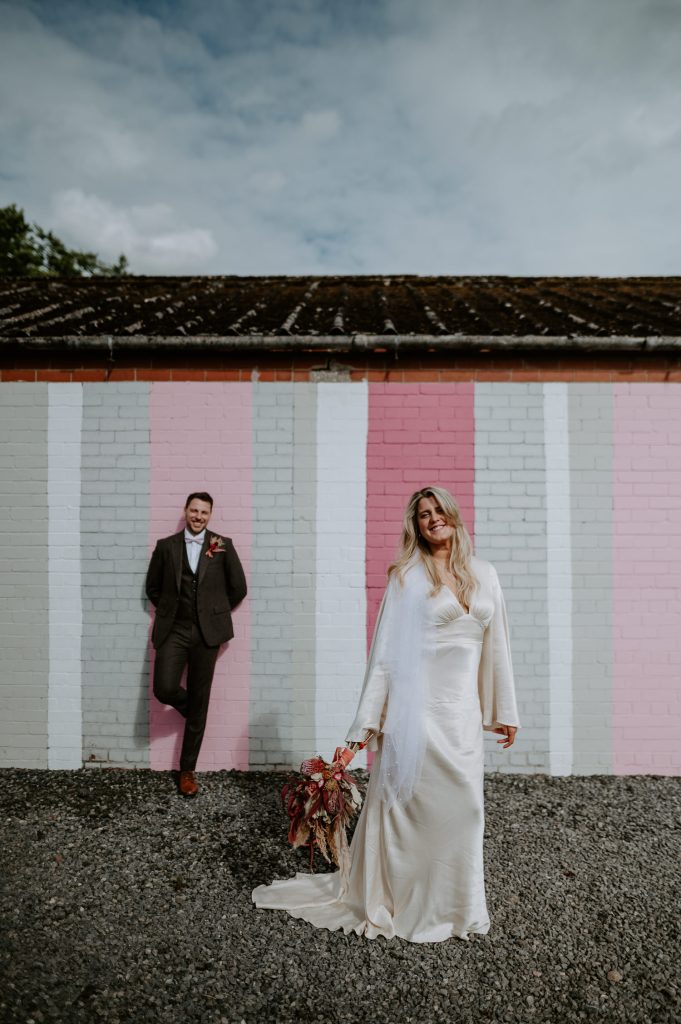 A bride stands in front of a groom in front of a pink brick wall.