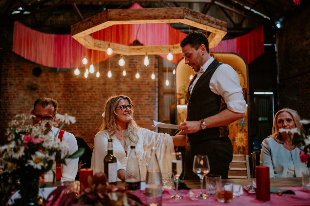 A groom delivers his speech whilst his bride smiles at his at White Syke Fields